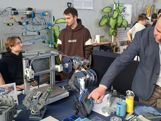 Mężczyzna w marynarce pokazuje studentom obsługę panelu sterowania nowoczesnego urządzenia technicznego w laboratorium. Obok nich znajdują się monitor z analizą obrazu, drukarka 3D oraz makieta silnika. Wszyscy są skupieni na praktycznej nauce.
