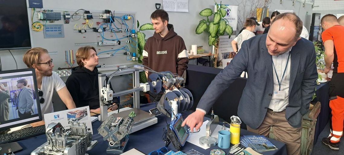 Mężczyzna w marynarce pokazuje studentom obsługę panelu sterowania nowoczesnego urządzenia technicznego w laboratorium. Obok nich znajdują się monitor z analizą obrazu, drukarka 3D oraz makieta silnika. Wszyscy są skupieni na praktycznej nauce.